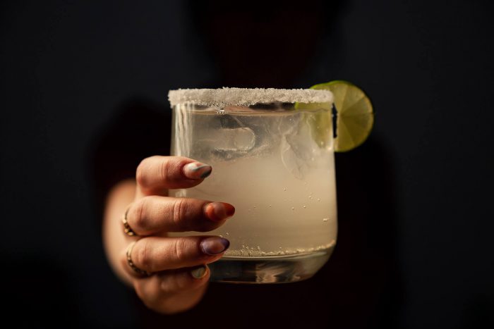 Hand holding margarita glass. The rim is salted and it has a lime.
