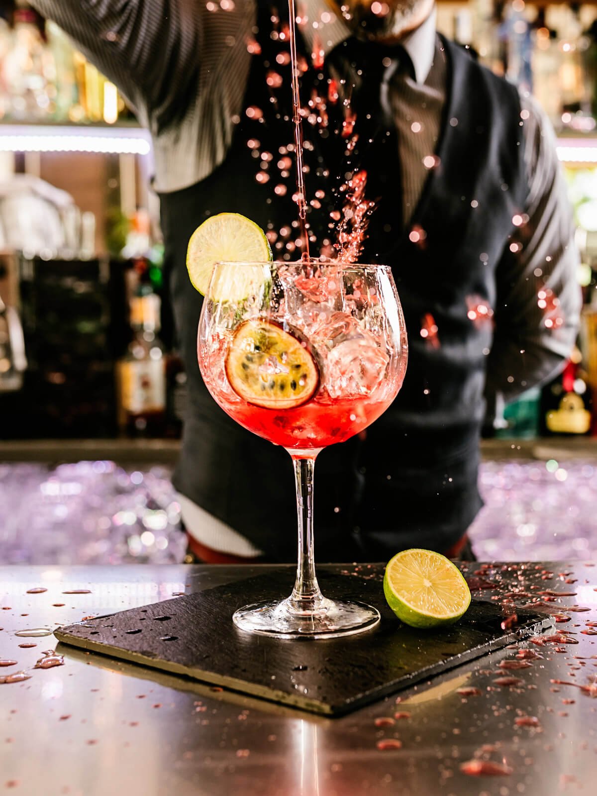 A fruity red drink sits on a bar while a bartender pours from above.