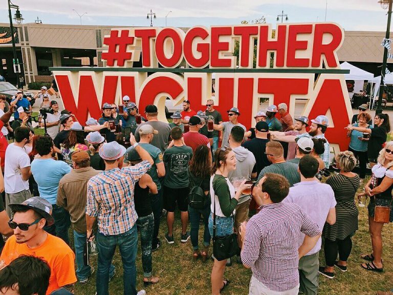Outdoor event with crowd standing in front of a giant red #TOGETHERWICHITA sign.