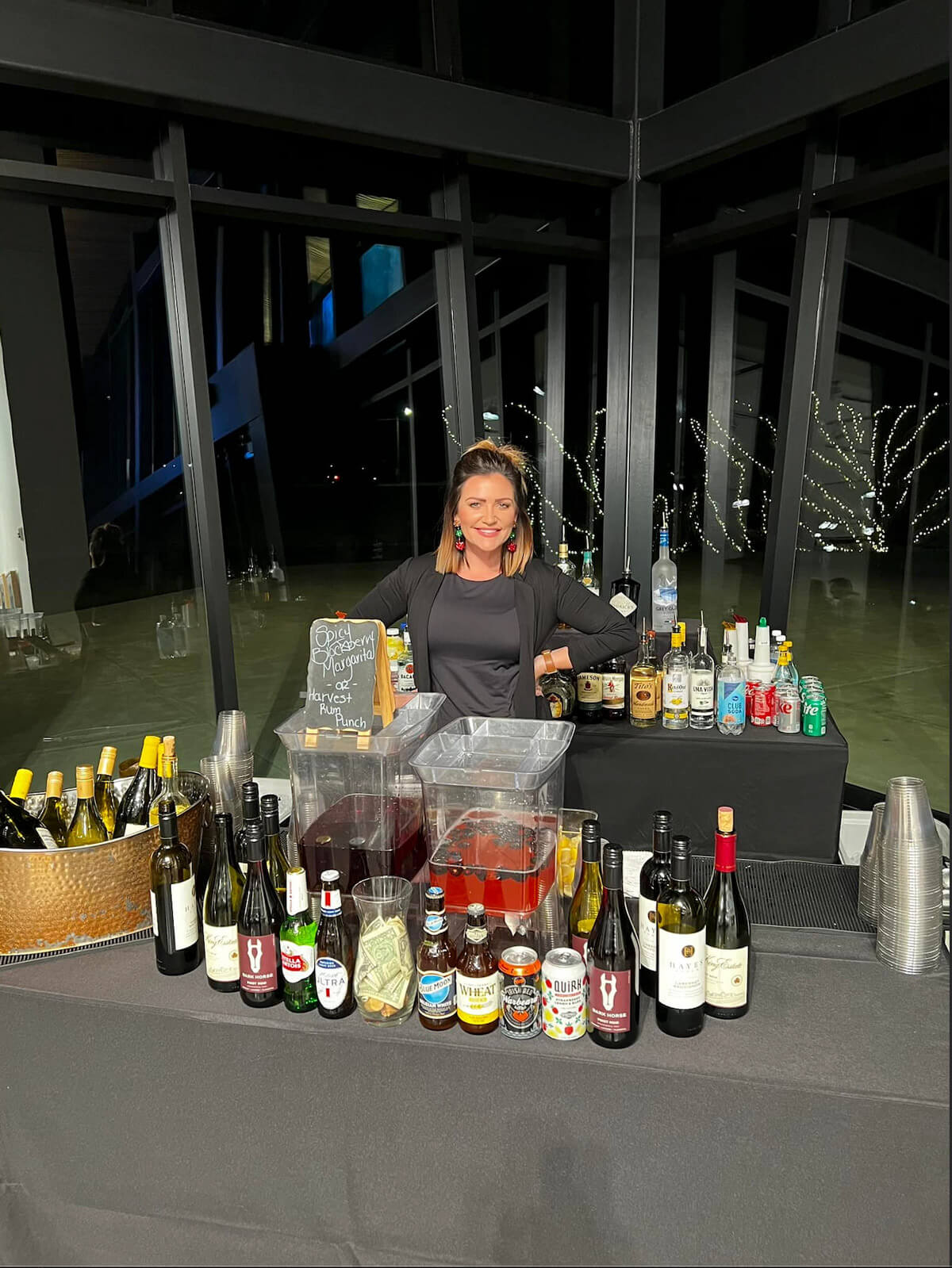 A smiling, female Xclusive bartender stands behind a full bar.